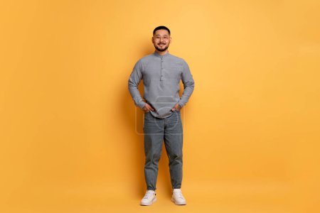 Photo for Smiling Young Asian Man Wearing Stylish Eyeglasses Posing Over Blue Studio Background, Handsome Millennial Guy Keeping Hands In Pockets And Looking At Camera, Full Length Shot With Copy Space - Royalty Free Image