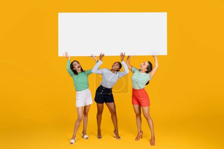 Photo for Happy multiracial young beautiful ladies in casual outfit carrying horizontal advertising board above their heads, blank space for advertisement or text, great sale season offer, orange background - Royalty Free Image