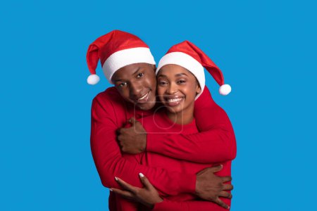 Téléchargez les photos : Vacances de Noël. Joyeux jeune couple noir en chapeaux rouges Santa étreignant sourire à la caméra posant sur fond bleu studio. Nouvel An, célébration de Noël et concept de saison d'hiver - en image libre de droit