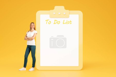 Photo for Cheerful confident adult european lady with crossed arms on chest near big plan with free space, do it list, isolated on yellow studio background. Recommendation future planning, motivation - Royalty Free Image