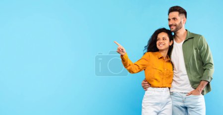 Foto de Gran anuncio y oferta. Feliz pareja europea abrazando, mujer apuntando al espacio vacío aislado sobre fondo azul, panorama. Venta en línea y promoción - Imagen libre de derechos
