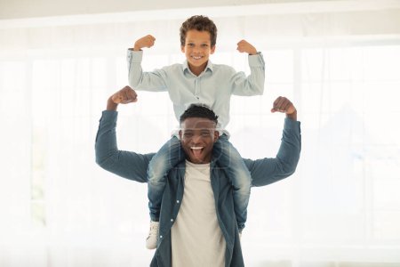 Foto de Somos fuertes. Papá afroamericano sosteniendo a su pequeño hijo en el cuello y mostrando sus bíceps, enseñando a su hijo a ser un líder en casa, demostrando fuerza. Criar al pequeño campeón Concepto - Imagen libre de derechos