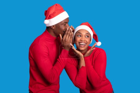 Photo for Secret Christmas Offer. Young Black Man Whispering Great News About Winter Sales To Excited Woman, Wearing Red Santa Hats Over Blue Studio Background. New Year And Xmas Holiday Season - Royalty Free Image