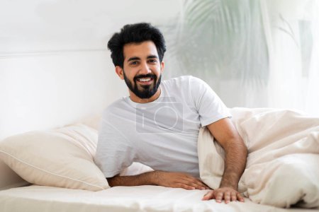 Téléchargez les photos : Portrait d'un jeune homme indien heureux se réveillant au lit le matin à la maison, gai Millénaire Oriental Guy reposant dans la chambre à coucher lumière et souriant à la caméra, bénéficiant du confort domestique, Copier l'espace - en image libre de droit