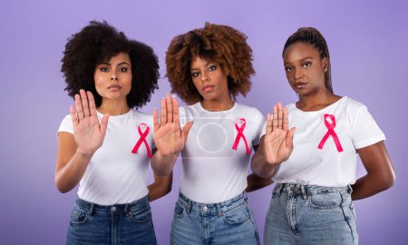 Téléchargez les photos : La sensibilisation au cancer du sein. Trois jeunes femmes noires gesticulant Stop To Camera, portant des t-shirts avec des rubans roses sur fond violet studio. Oncologie Maladie, concept du groupe de soutien - en image libre de droit