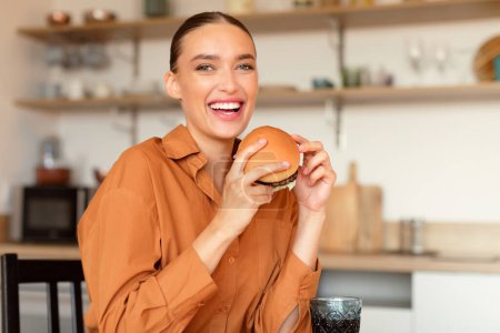 Foto de Retrato de mujer feliz sosteniendo gran hamburguesa sabrosa en las manos y sonriendo a la cámara, sentado en la mesa en la cocina, espacio libre. Comedor de comida chatarra hembra mostrando hamburguesa casera - Imagen libre de derechos