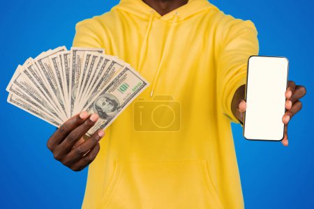 Téléchargez les photos : Jeune homme noir montre beaucoup d'argent dollars et smartphone avec écran blanc, isolé sur fond bleu, studio. Bénéfice, victoire et richesse, application financière, recommandation bancaire - en image libre de droit