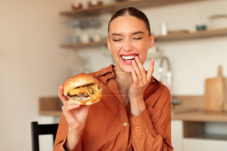 Foto de La gula. Mujer feliz comiendo hamburguesa casera, sosteniendo sabrosa comida rápida y sonriendo, sentado en el interior de la cocina, espacio libre. Nutrición poco saludable y hábito de atracones - Imagen libre de derechos