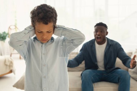 Photo for Domestic Violence Against Children. Mad African American Dad Shouting At Scared Kid Son While Child Covering Ears At Home. Selective Focus On Unhappy Little Boy During Quarrel With Father - Royalty Free Image
