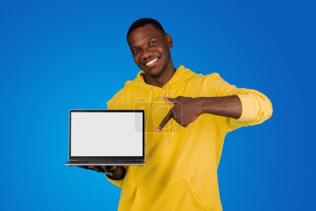 Téléchargez les photos : Un jeune homme noir souriant pointe du doigt un ordinateur portable avec écran vide, isolé sur fond bleu, studio. Recommandation pour le travail et l'étude à distance avec l'appareil, site Web, blog - en image libre de droit