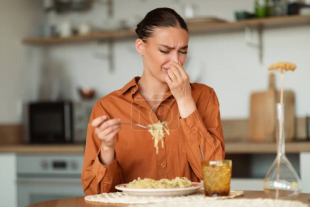 Photo for Awful delivery food. Young woman closing nose while eating spaghetti, lady looking at plate, sitting at table in kitchen interior. Foul food concept - Royalty Free Image