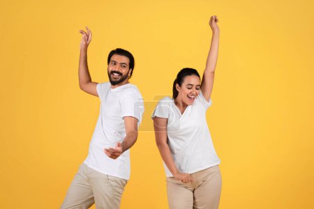 Photo for Cheerful millennial arab guy and woman dance, celebrating victory, raising hands, making sign of success, isolated on yellow studio background. Spare time, fun and relationships - Royalty Free Image