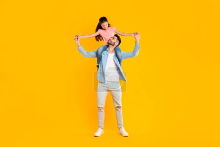Photo for Happy moments. Excited man riding his daughter on shoulders, having fun together, father holding child hands and smiling at each other over yellow background, full length - Royalty Free Image