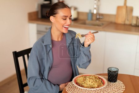 Foto de Mujer embarazada joven satisfecha disfrutar de comer espaguetis caseros cocinados, sentado en la mesa en la cocina, degustación de pasta y sonriendo, interior del hogar, espacio para copiar - Imagen libre de derechos