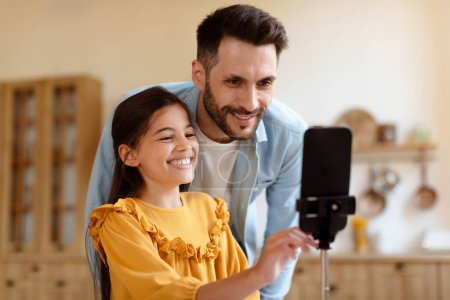 Photo for Online Fun With Dad. Happy Preteen Kid Daughter And Her Father Using Smartphone On Tripod, Shooting Video For Blog Or Making Photo Via Phone, Having Fun With Modern Gadget At Home. Selective Focus - Royalty Free Image