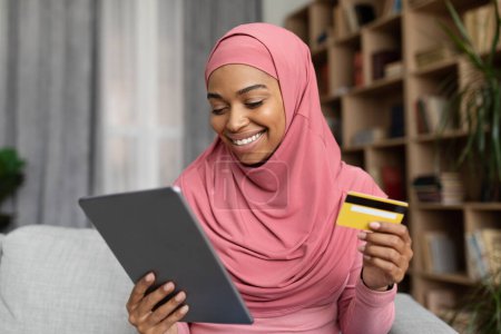 Foto de Banca fácil virtual. Feliz joven musulmana negra en hijab sentada en el sofá, sosteniendo la tableta digital y la tarjeta bancaria, banca en línea en casa, espacio para copiar - Imagen libre de derechos