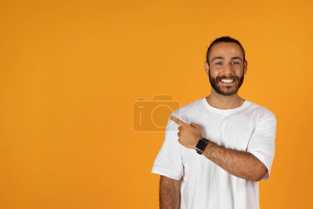 Photo for Glad millennial caucasian guy with beard in white t-shirt point finger at copy space, isolated on orange studio background. Recommendation ad and offer, professional advice to sale, work, study - Royalty Free Image