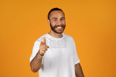 Téléchargez les photos : Heureux homme caucasien millénaire avec barbe en t-shirt blanc pointe doigt à la caméra, isolé sur fond de studio orange. Confiance, geste de motivation, recommandation, votre choix, votre tour - en image libre de droit