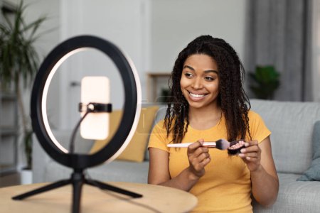 Photo for Beauty blog. Black lady blogger advertising new cosmetics products, making makeup filming on cellphone set on ring lamp, living room. Professional blogging concept - Royalty Free Image