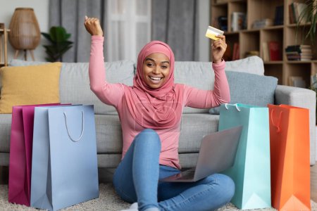 Téléchargez les photos : Femme musulmane noire excitée shopaholic en hijab assis avec beaucoup de sacs à provisions colorés, en utilisant un ordinateur portable et une carte de crédit à la maison, secouant les poings. E-commerce, concept d'achat en ligne réussi - en image libre de droit