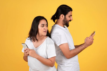 Photo for Shocked millennial arab woman looking at husband phone, isolated on yellow studio background. Online cheating, chat, family and relationship problems, mistrust, gadget addiction - Royalty Free Image