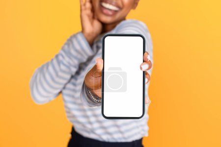 Téléchargez les photos : Méconnaissable souriant jeune femme noire montrant smartphone avec écran blanc vide maquette espace vide pour la publicité ou le texte, fond jaune. Application mobile, concept d'offre en ligne - en image libre de droit