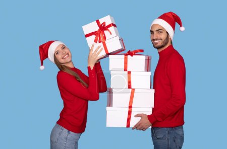 Foto de Pareja joven feliz en las cajas de apilamiento de los sombreros de Santa con los regalos de Navidad y sonriendo en la cámara, hombre alegre y mujer sosteniendo regalos, celebrando días festivos de Navidad, posando en fondo azul del estudio - Imagen libre de derechos