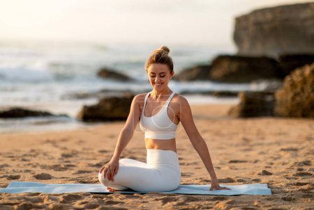Téléchargez les photos : Jeune athlète européenne en tenue de sport assis en position lotus pratiquant le yoga, profitant d'exercices de corps étirant sur la plage de la mer. S'adapter à l'extérieur, le sport et la perte de poids - en image libre de droit