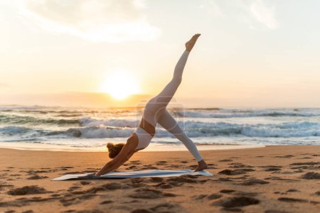 Téléchargez les photos : Jeune femme en vêtements de sport faisant de l'exercice de yoga avec la jambe levée à l'extérieur, ayant l'entraînement sur la plage de la mer au coucher du soleil, vue latérale, pleine longueur. Dame qui s'étire dehors - en image libre de droit