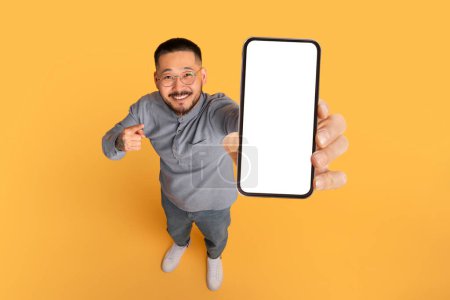 Photo for Check This. Smiling Asian Man Pointing At Big Blank Smartphone In His Hand, Happy Millennial Guy Advertising Online Offer Or Mobile App, Standing Over Yellow Studio Background, Collage, Mockup - Royalty Free Image