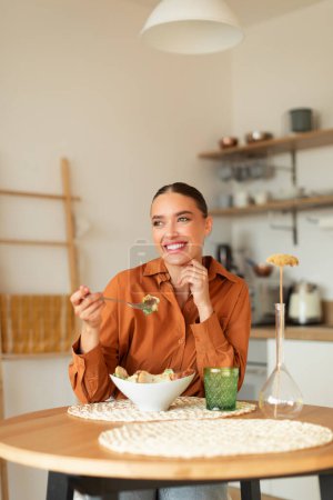 Photo for Cheerful dreamy woman enjoying delicious caesar salad, sitting at table in kitchen interior, looking aside and smiling, vertical shot, cropped, copy space - Royalty Free Image