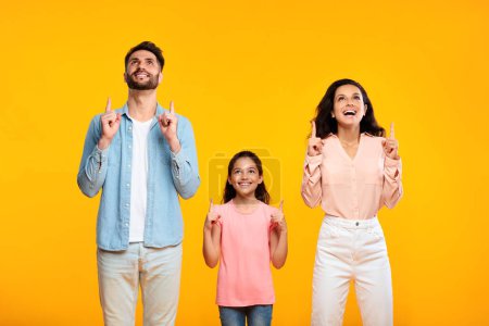 Foto de Familia europea alegre de tres dedos mirando y señalando hacia arriba, posando de pie sobre el fondo amarillo, espacio libre. Padres y niñas mostrando lugar para el anuncio por encima de la cabeza - Imagen libre de derechos
