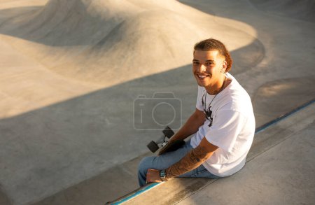 Foto de Happy guy skateboarder en camiseta descansando después de practicar trucos, sentado en la rampa de hormigón y sonriendo a la cámara, por encima de la vista, espacio libre - Imagen libre de derechos