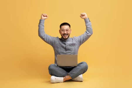 Téléchargez les photos : Gagner en ligne. Joyeux jeune homme asiatique célébrant le succès avec ordinateur portable, gai Millennial Guy assis sur le sol sur fond jaune studio et levant les poings, acclamant chance et victoire, Copier l'espace - en image libre de droit