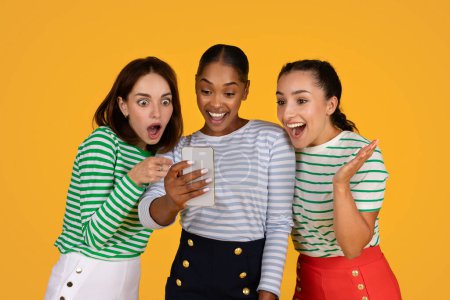 Photo for Season sale, black friday deal, e-commerce. Excited attractive millennial multiethnic ladies shopaholics looking at smartphone screen, exclaiming and gesturing, isolated on yellow background - Royalty Free Image