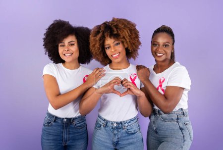 Photo for Oncology Awareness. Trio Of Positive African American Ladies With Pink Breast Cancer Ribbons Hugging, Woman Shaping Fingers Like Heart Over Purple Studio Background. Female Support Group - Royalty Free Image
