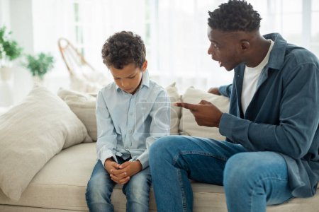 Photo for Child Behavior Problem. Angry Black Dad Scolding Unhappy Kid Son, Shouting And Pointing Finger At Boy Sitting On Sofa At Home. Aggressive Father Issue, Domestic Violence Concept. Selective Focus - Royalty Free Image