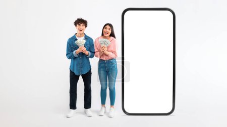 Photo for Great Luck. Joyful Young Couple Holding Money Cash Near Huge Cellphone With Blank Touchscreen, Advertising Online Cashback Service Or Financial Application For Phone, White Background, Panorama - Royalty Free Image