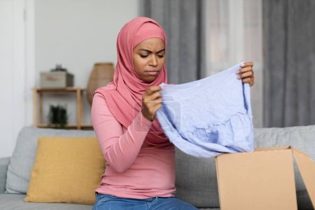 Téléchargez les photos : Femme musulmane noire déçue cochant la case postale, ouvrant la livraison et vérifiant les vêtements, ayant une expression faciale négative, assise sur un canapé à la maison. Concept de livraison - en image libre de droit