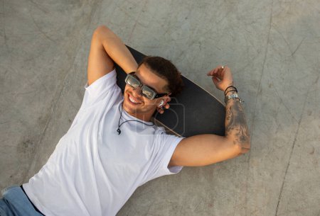 Photo for Stylish tattooed man relaxing on skateboard and listening music in earpads, having rest after skateboarding exercises, top view, free space. Guy relaxing in skate park - Royalty Free Image