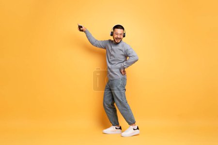 Téléchargez les photos : Heureux homme asiatique portant des écouteurs sans fil dansant sur fond jaune studio, gai Millennial Guy écouter de la musique et s'amuser, profiter de chansons préférées, pleine longueur tourné avec de l'espace de copie - en image libre de droit