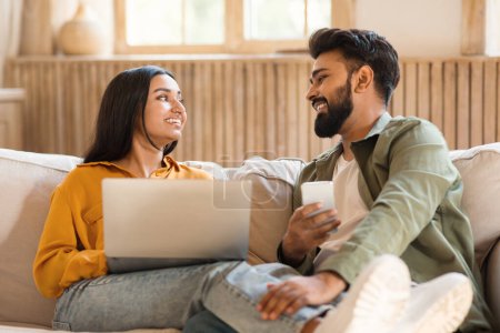 Téléchargez les photos : Week-end passe-temps concept. Heureux couple indien utilisant ordinateur portable et smartphone à la maison, se regardant tout en se relaxant sur le canapé. Conjoints naviguant sur Internet - en image libre de droit