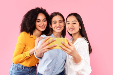 Foto de Feliz multirracial amigas haciendo selfie en el teléfono celular, tomando fotos para las redes sociales, de pie juntos sobre fondo de estudio rosa. concepto de amistad femenina - Imagen libre de derechos