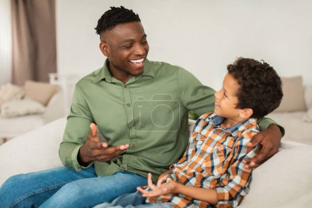 Foto de Tiempo de calidad con papá. Happy Black Father y Little Son comparten sus historias hablando y disfrutando de la conversación, sentados abrazándose en el sofá en casa. Relaciones familiares y comunicación - Imagen libre de derechos