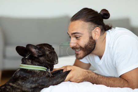 Téléchargez les photos : Joyeux jeune homme jouant avec le chien bouledogue français dans le lit le matin. Millennial guy se réveille et chien animal de compagnie debout à côté du lit. Concept d'amitié entre l'homme et l'animal - en image libre de droit