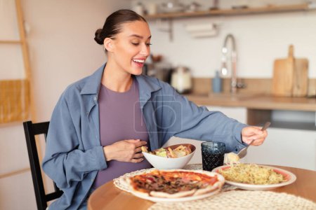 Téléchargez les photos : Femme enceinte caucasienne affamée au choix parmi la pizza, la salade et manger des pâtes, ayant le désir de malbouffe, assis à table à l'intérieur de la cuisine, espace libre - en image libre de droit