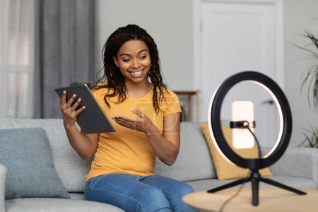 Photo for Modern work and hobby. Positive black female blogger shooting video blog, showing tablet, broadcasting in living room interior. Review on modern technology, ad and offer - Royalty Free Image