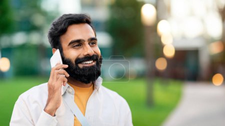Foto de Retrato al aire libre del indio llamando y hablando por teléfono mirando hacia un lado, parado afuera en el área urbana de la ciudad, caminando en una calle por la noche y comunicándose por teléfono celular. Panorama - Imagen libre de derechos