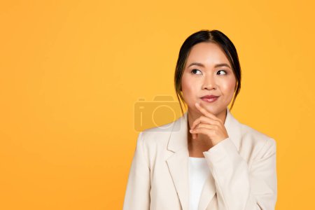 Téléchargez les photos : Pensive positive jeune femme asiatique en costume pense, en regardant l'espace vide, isolé sur fond de studio jaune. Choix, émotions des gens, annonce et offre, solution et idée créent - en image libre de droit