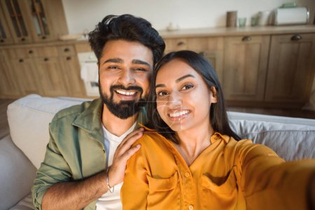 Foto de Retrato de cerca de una alegre pareja india tomando selfie, descansando en el sofá en casa y sonriendo, hombre y mujer orientales abrazándose, posando en el interior de la sala de estar - Imagen libre de derechos